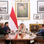 Group of officials sit around a polished wooden conference table in a formal meeting with the indonesian flag behind them and framed portraits on the wall behind the center speaker
